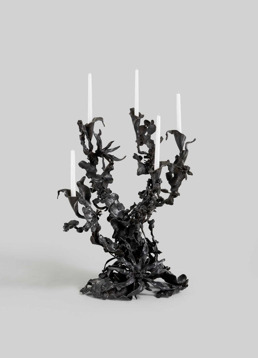 The VIYA Gardens Of India Candelabra Small is a sculptural black metal piece with five white candles, featuring twisted, organic, leafy forms that evoke the intricate beauty of an Indian garden. Set against a plain light gray background.