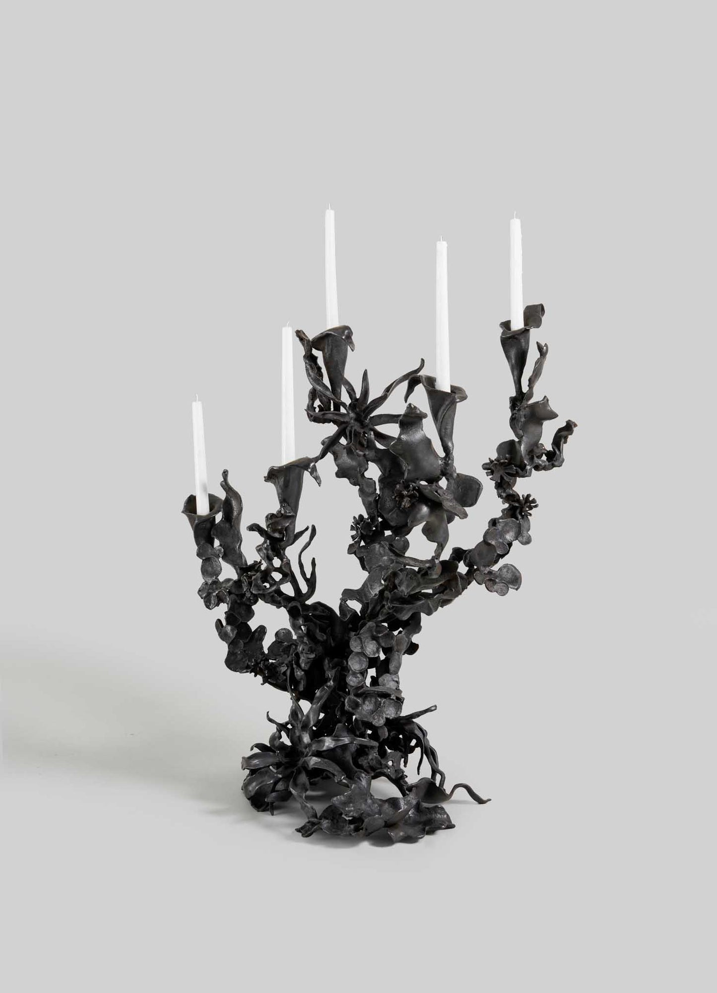 The VIYA Gardens Of India Candelabra Small is an ornate black metal piece with five branches for white candles, featuring intricate, blossom-inspired details that evoke the elegance of an Indian garden against a plain gray background.