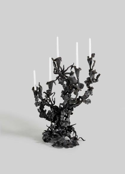 The VIYA Gardens Of India Candelabra Small is an ornate black metal piece with five branches for white candles, featuring intricate, blossom-inspired details that evoke the elegance of an Indian garden against a plain gray background.
