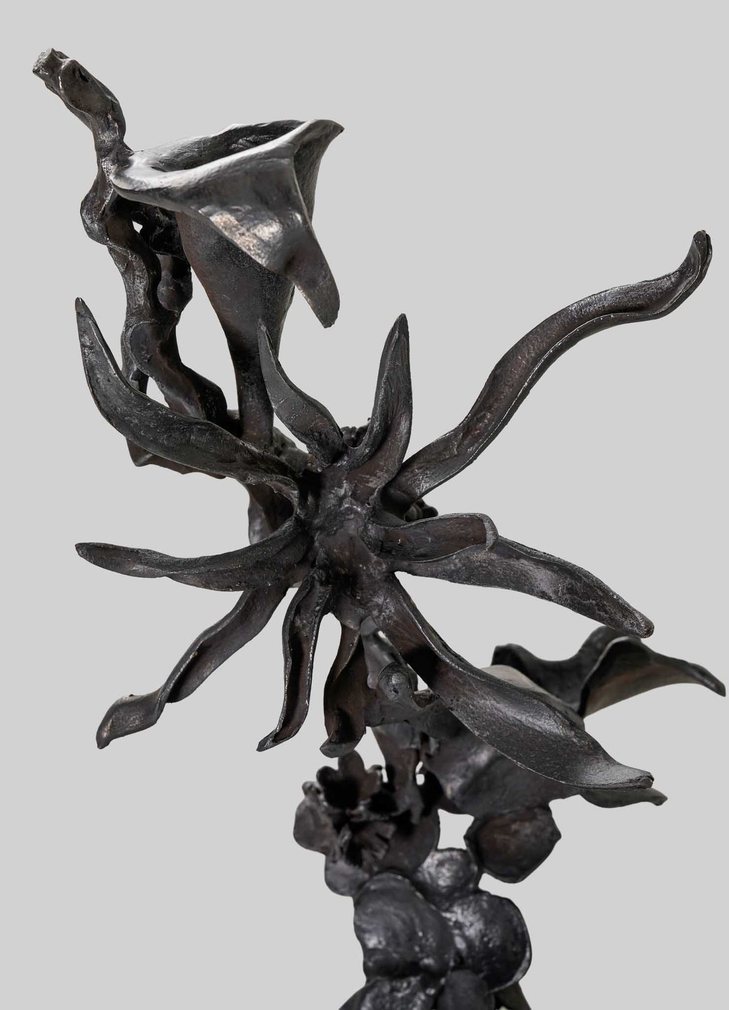 The VIYA Gardens Of India Candelabra Small features abstract dark metal with twisted, elongated forms reminiscent of petals and stems, evoking fragrant blossoms from an Indian garden against a plain light gray background.