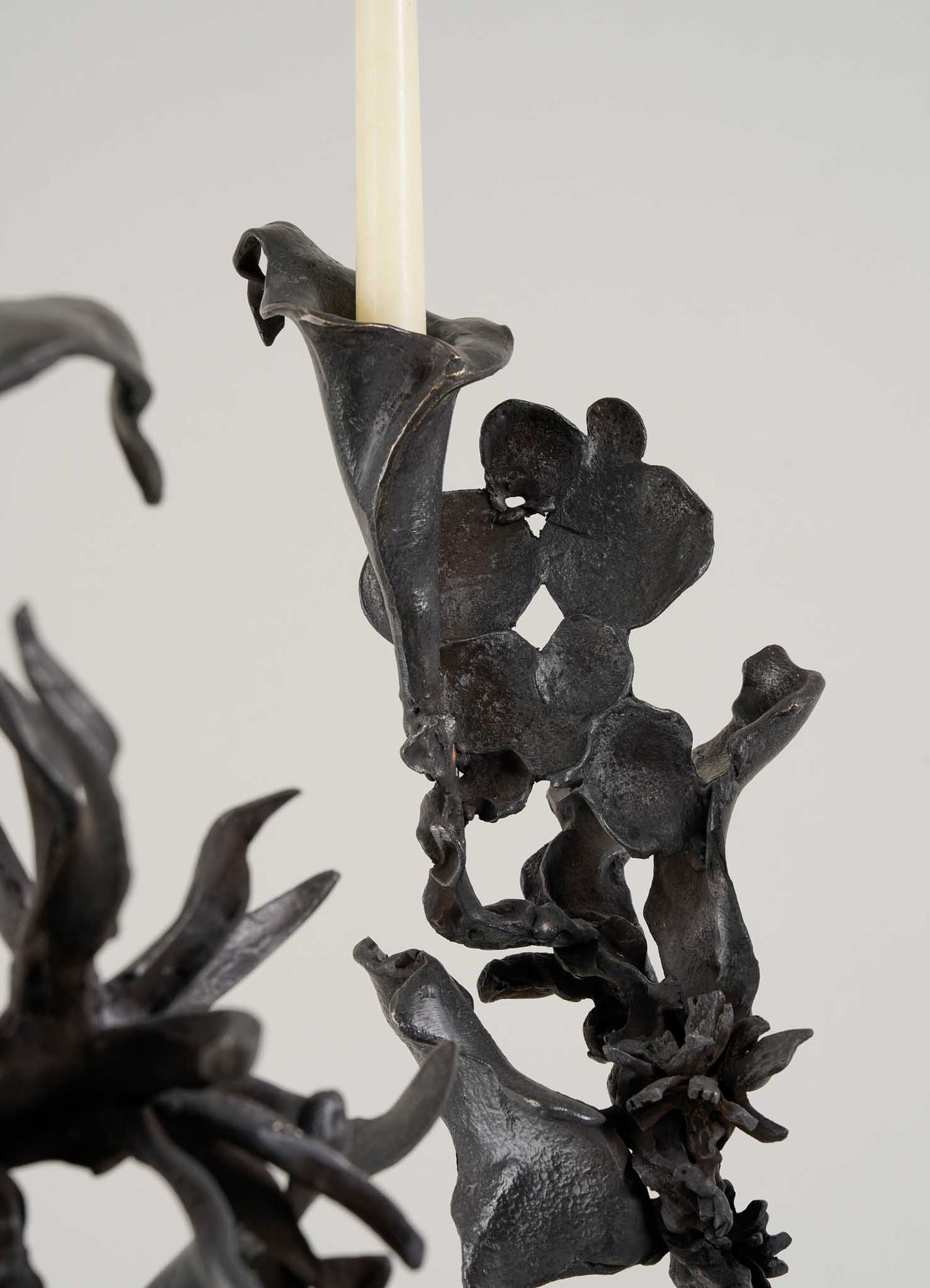 Close-up of the Gardens Of India Candelabra Small by VIYA, a black metal sculpture with abstract, leaf- and flower-like shapes holding a slender candle aloft against a plain light background.