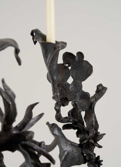 Close-up of the Gardens Of India Candelabra Small by VIYA, a black metal sculpture with abstract, leaf- and flower-like shapes holding a slender candle aloft against a plain light background.