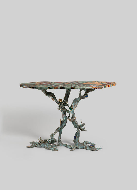 The VIYA Glory Lily Multicolour Console Table features a vibrant mosaic-style top and a sculptural cast brass base inspired by twisting tree branches and the Glory Lily flower, set against a plain light gray background.