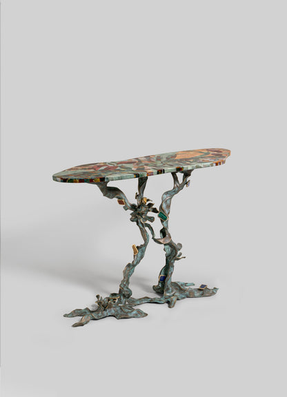 The VIYA Glory Lily Multicolour Console Table features an oval mosaic top and sculpted cast brass legs inspired by the graceful Glory Lily flower, set against a plain light gray background.