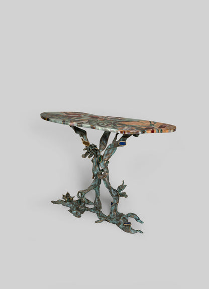 The VIYA Glory Lily Multicolour Console Table features a colorful, irregular top and a sculptural cast brass base inspired by Glory Lily roots, displayed on a plain light gray background.