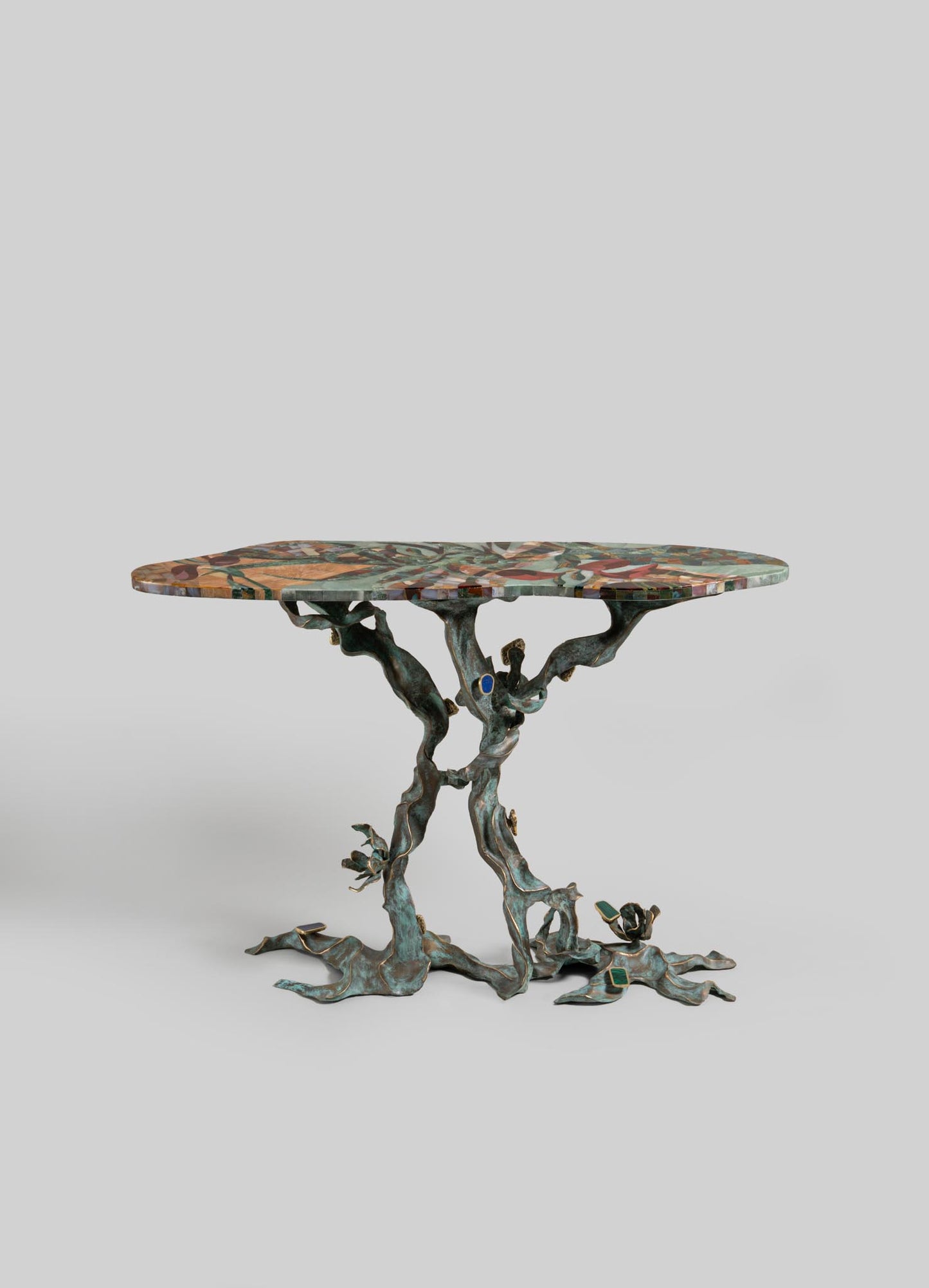 The VIYA Glory Lily Multicolour Console Table features a vibrant mosaic-patterned oval top and a sculpted cast brass base reminiscent of twisted tree branches, inspired by the Glory Lily flower, shown against a plain light gray background.