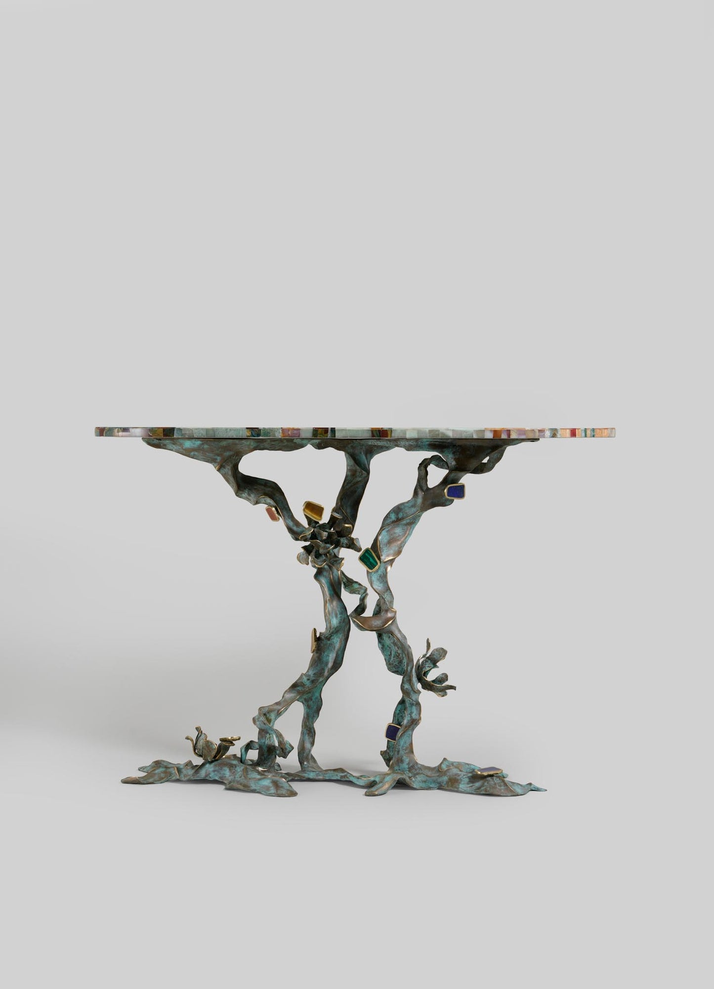 The VIYA Glory Lily Multicolour Console Table features a vibrant, floral-patterned top inspired by the Glory Lily and a sculptural cast brass base with intertwined branch design, shown against a plain light gray background.