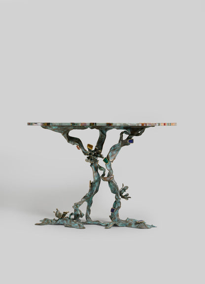 The VIYA Glory Lily Multicolour Console Table features a vibrant, floral-patterned top inspired by the Glory Lily and a sculptural cast brass base with intertwined branch design, shown against a plain light gray background.