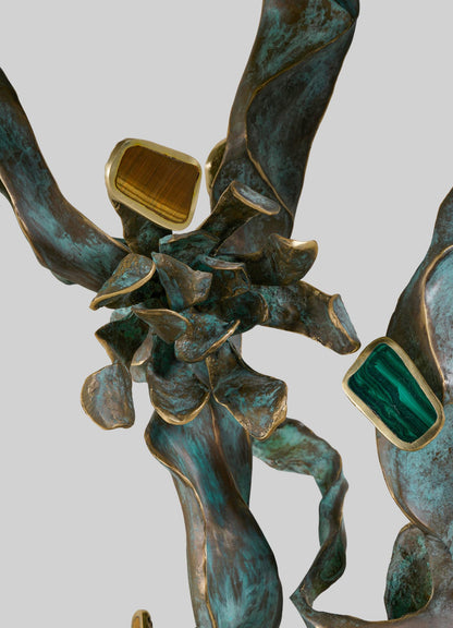 The VIYA Glory Lily Multicolour Console Table features jagged, twisting bronze forms with a weathered green patina, decorative rectangular insets of brown and green stones edged in gold, all set against a light gray background.