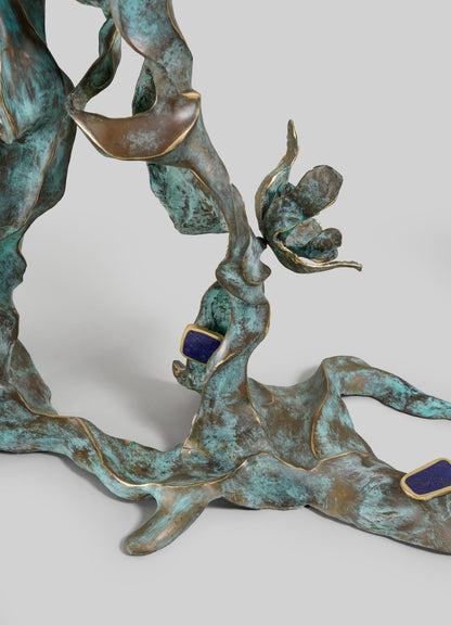 The VIYA Glory Lily Multicolour Console Table features organic, twisting forms inspired by the Glory Lily flower, a bronze and green patina finish, gold accents, and two small rectangular blue elements on a plain gray background.