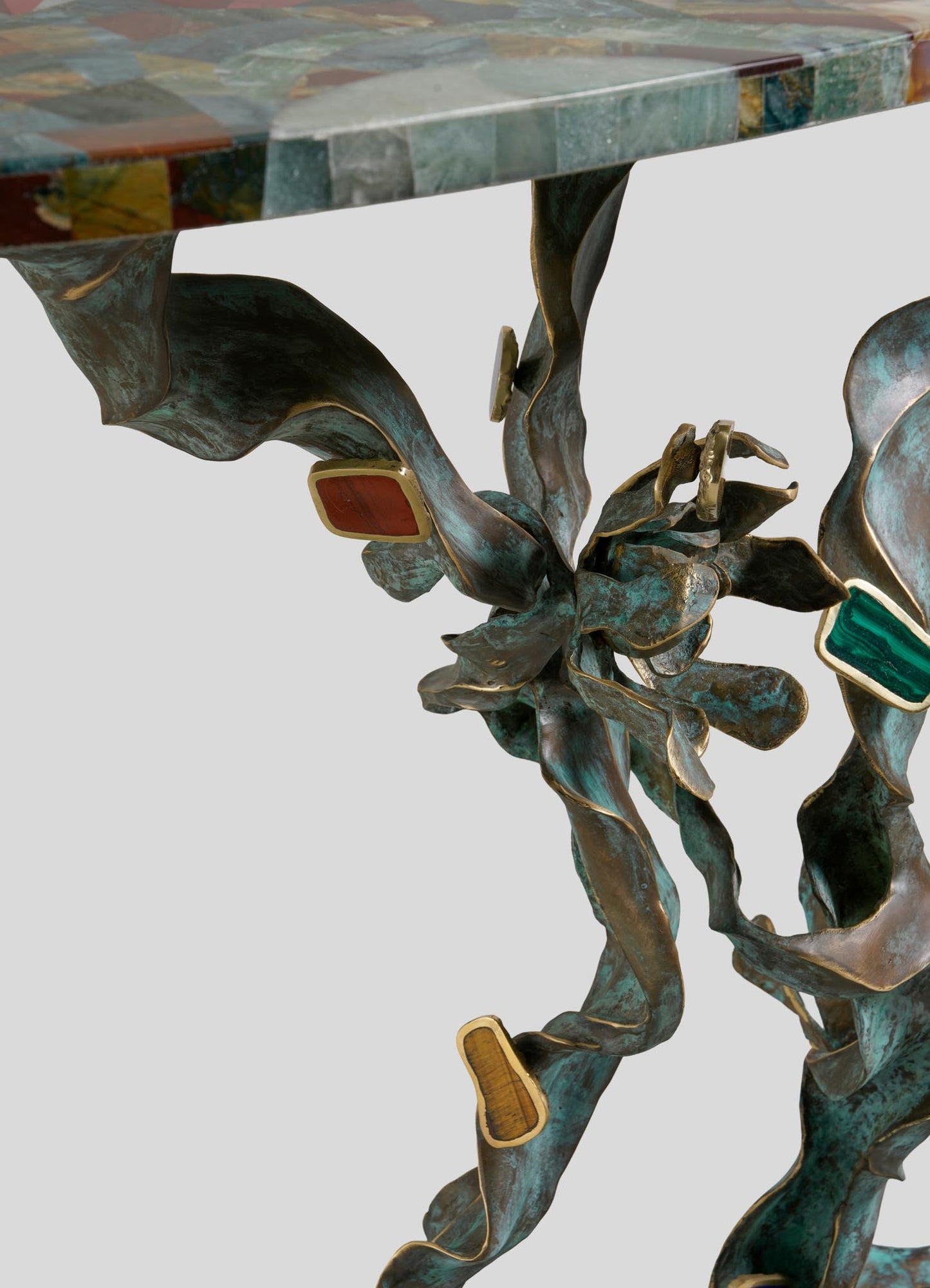 A close-up of the VIYA Glory Lily Multicolour Console Table, showcasing its textured, twisting cast brass base with colorful stone inlays and a polished stone top. The backdrop is plain and light.