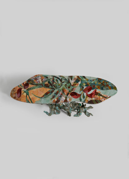 The VIYA Glory Lily Multicolour Console Table features a vibrant, abstract-patterned oval top set on an intricate sculptural cast brass base shaped like intertwined branches, inspired by the Glory Lily flower, shown against a light gray background.