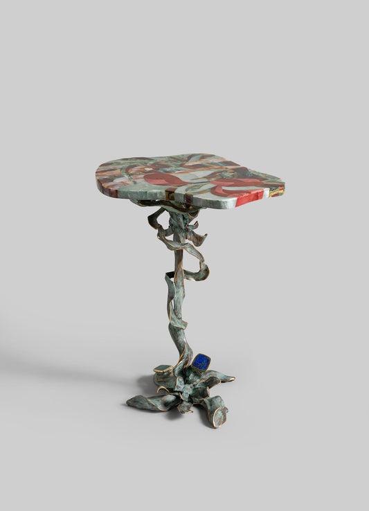 The Glory Lily Side Table by VIYA features a colorful, uniquely shaped top and a twisting cast brass base inspired by the Glory Lily roots, all set against a plain gray background.