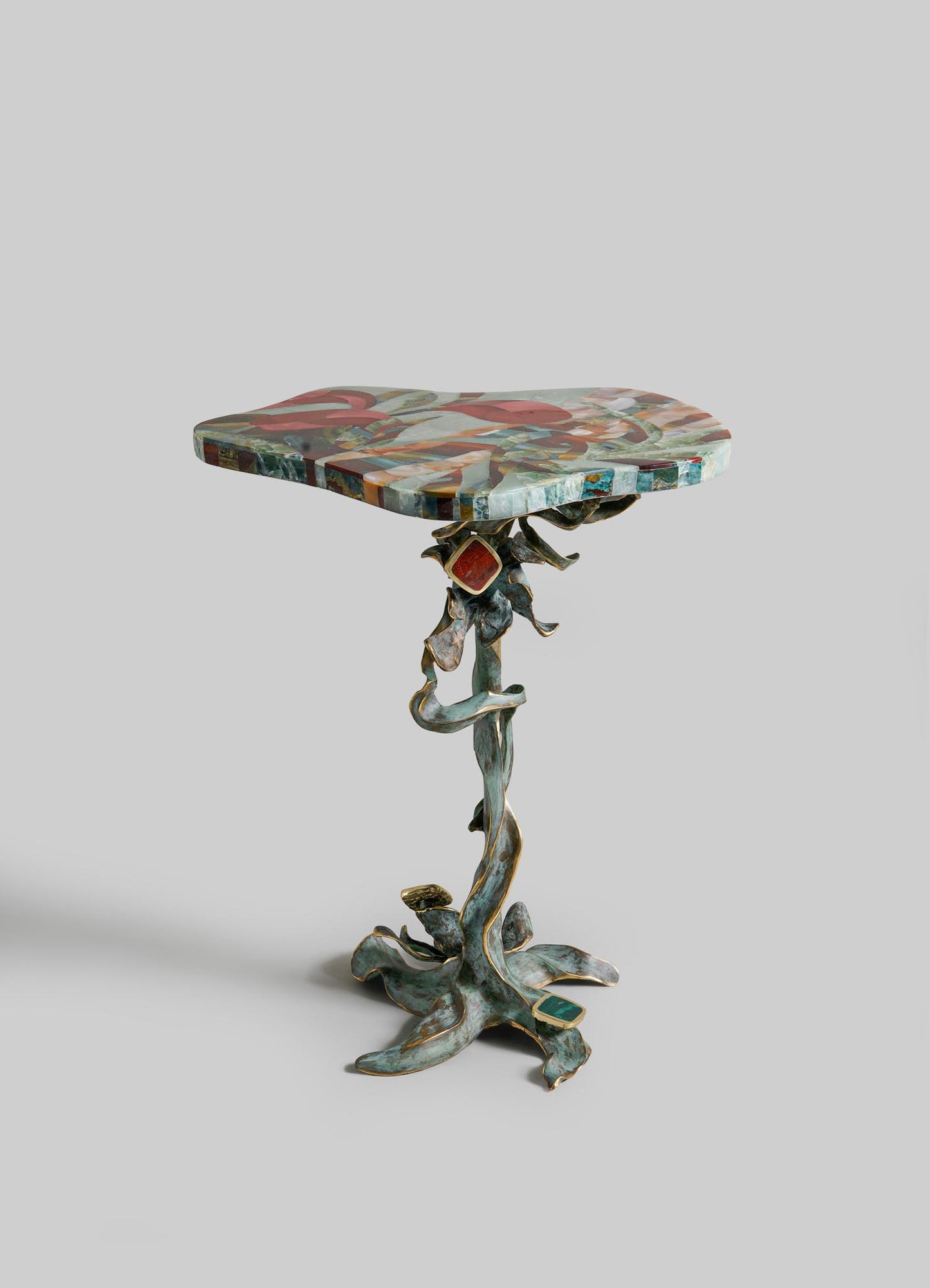 The VIYA Glory Lily Multicolour Side Table Large features a vibrant, irregular top and a twisted cast brass base inspired by the Glory Lily flower, resembling intricate intertwined vines, shown against a plain light-gray background.