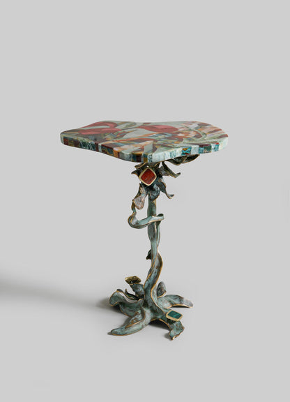 The VIYA Glory Lily Multicolour Side Table Large features a vibrant, irregular top and a twisted cast brass base inspired by the Glory Lily flower, resembling intricate intertwined vines, shown against a plain light-gray background.