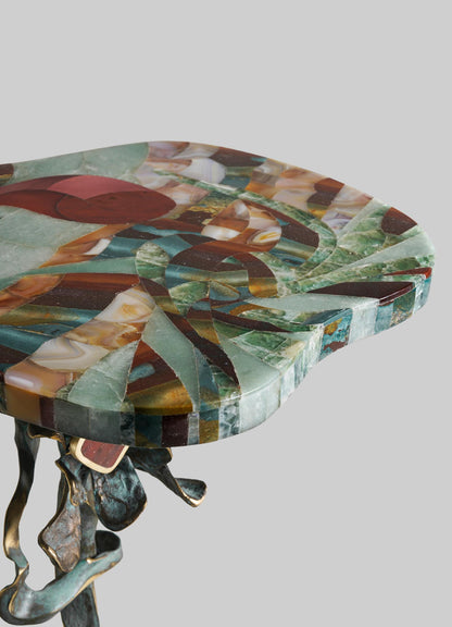The VIYA Glory Lily Multicolour Side Table Large features a polished, abstract-patterned stone top, a sculpted cast brass base with subtle Glory Lily motifs, shown against a plain gray background.