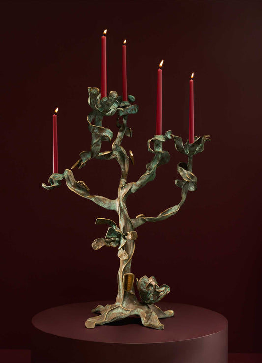 The VIYA Glory Lily Candelabra, inspired by the elegant Glory Lily flower, features sculptural cast brass branches holding five red candles, shown on a round maroon pedestal against a deep maroon background.
