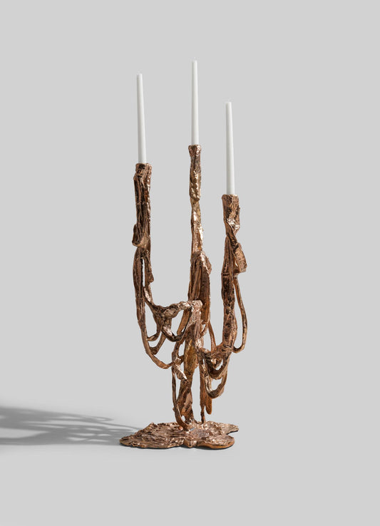 The VIYA Golden Banyan Candelabra is a sculptural bronze piece with pleated, melted-look details and three holders for white taper candles, shown unlit against a plain light grey background.