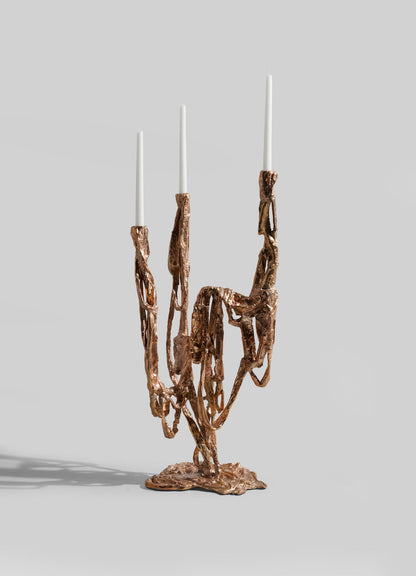 The VIYA Golden Banyan Candelabra features three arms with an abstract, organic bronze design and chain-like details. Each arm holds a tall white candle, set against a light gray background.
