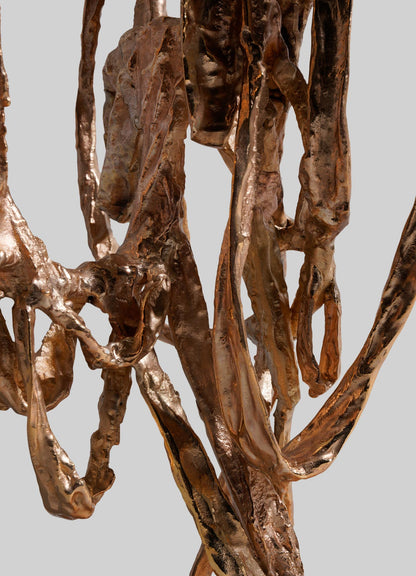 Close-up of VIYA’s Golden Banyan Candelabra: twisted, ribbon-like forms with a shiny, reflective surface evoke flowing pleated fabric art. The organic, elegant bronze sculpture stands out against a plain light gray background.