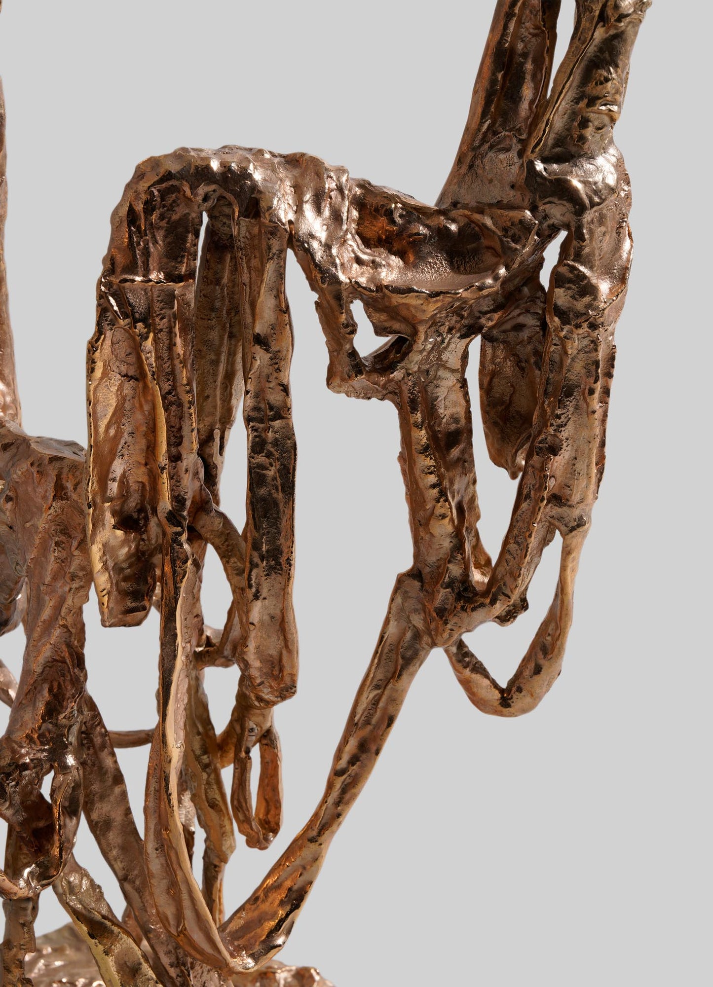Close-up of the VIYA Golden Banyan Candelabra, featuring twisted and looping bronze metal strips reminiscent of pleated fabric art, set against a plain grey background.