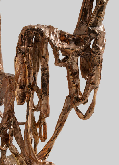 Close-up of the VIYA Golden Banyan Candelabra, featuring twisted and looping bronze metal strips reminiscent of pleated fabric art, set against a plain grey background.