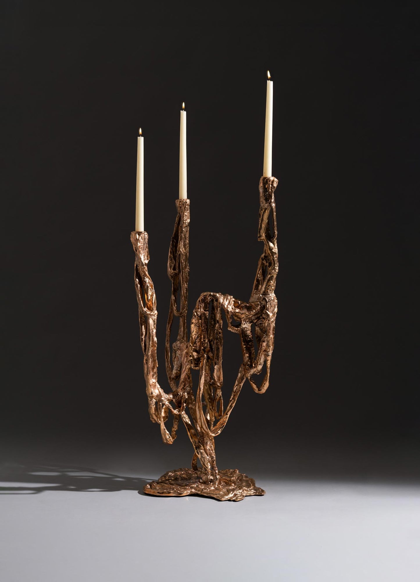 The VIYA Golden Banyan Candelabra, inspired by pleated fabric art, features sculptural bronze forms supporting three tall white candles and creates dramatic shadows on a gray surface against a dark background.