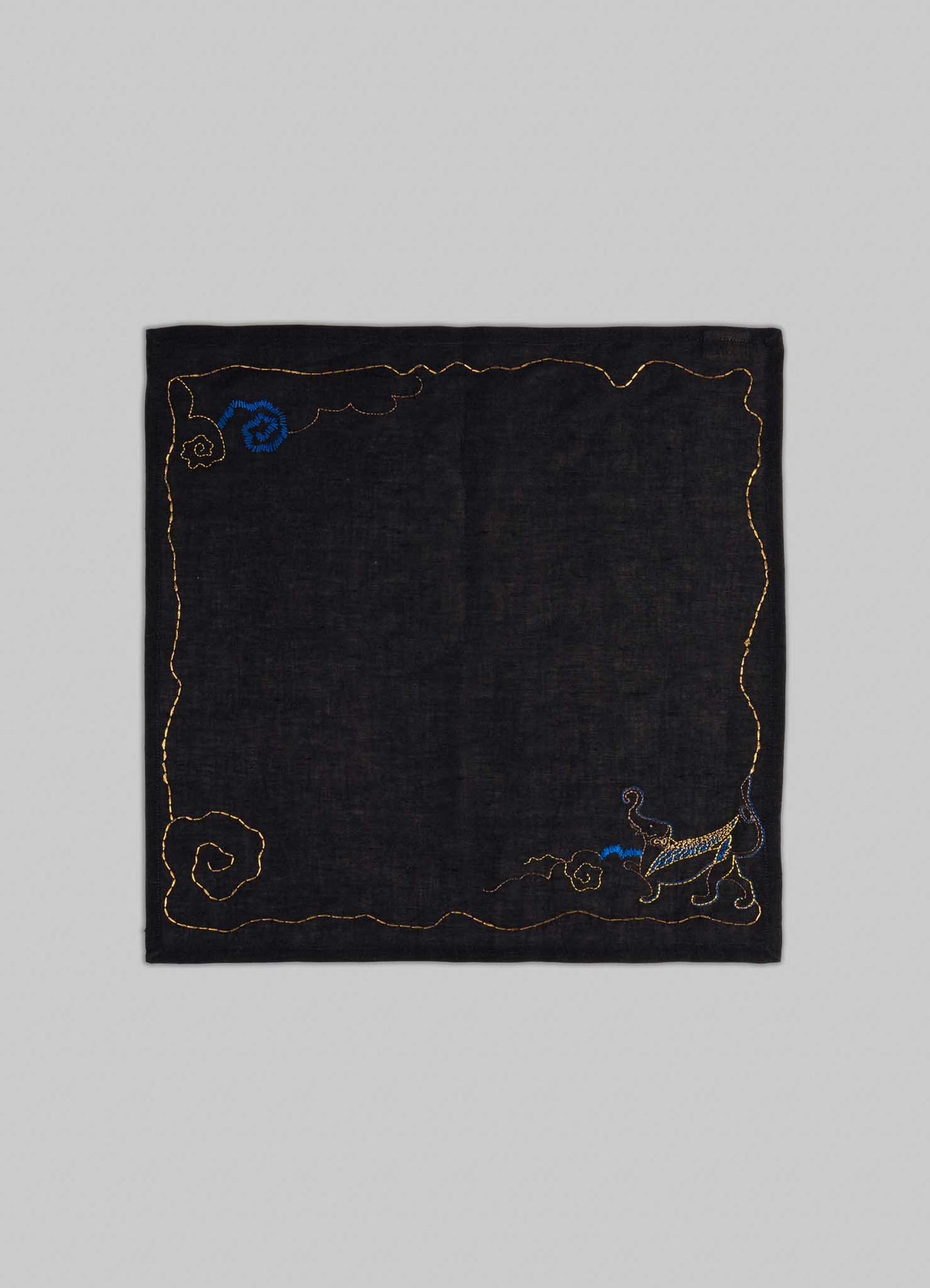 The Gajasimha Hand Embroidered Black & Gold Napkin by Viya features a gold-stitched black square design with spiral patterns and a blue-gold cat in the lower right, inspired by ancient Indian art and the mythical guardian Gajasimha.