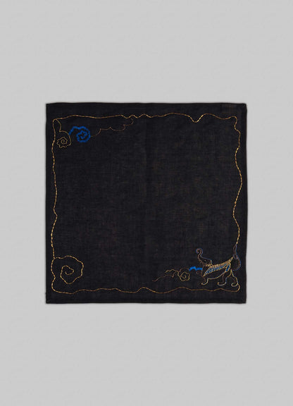 The Gajasimha Hand Embroidered Black & Gold Napkin by Viya features a gold-stitched black square design with spiral patterns and a blue-gold cat in the lower right, inspired by ancient Indian art and the mythical guardian Gajasimha.