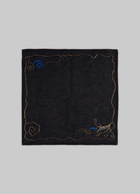 The Gajasimha Hand Embroidered Black & Gold Napkin by Viya features a gold-stitched black square design with spiral patterns and a blue-gold cat in the lower right, inspired by ancient Indian art and the mythical guardian Gajasimha.