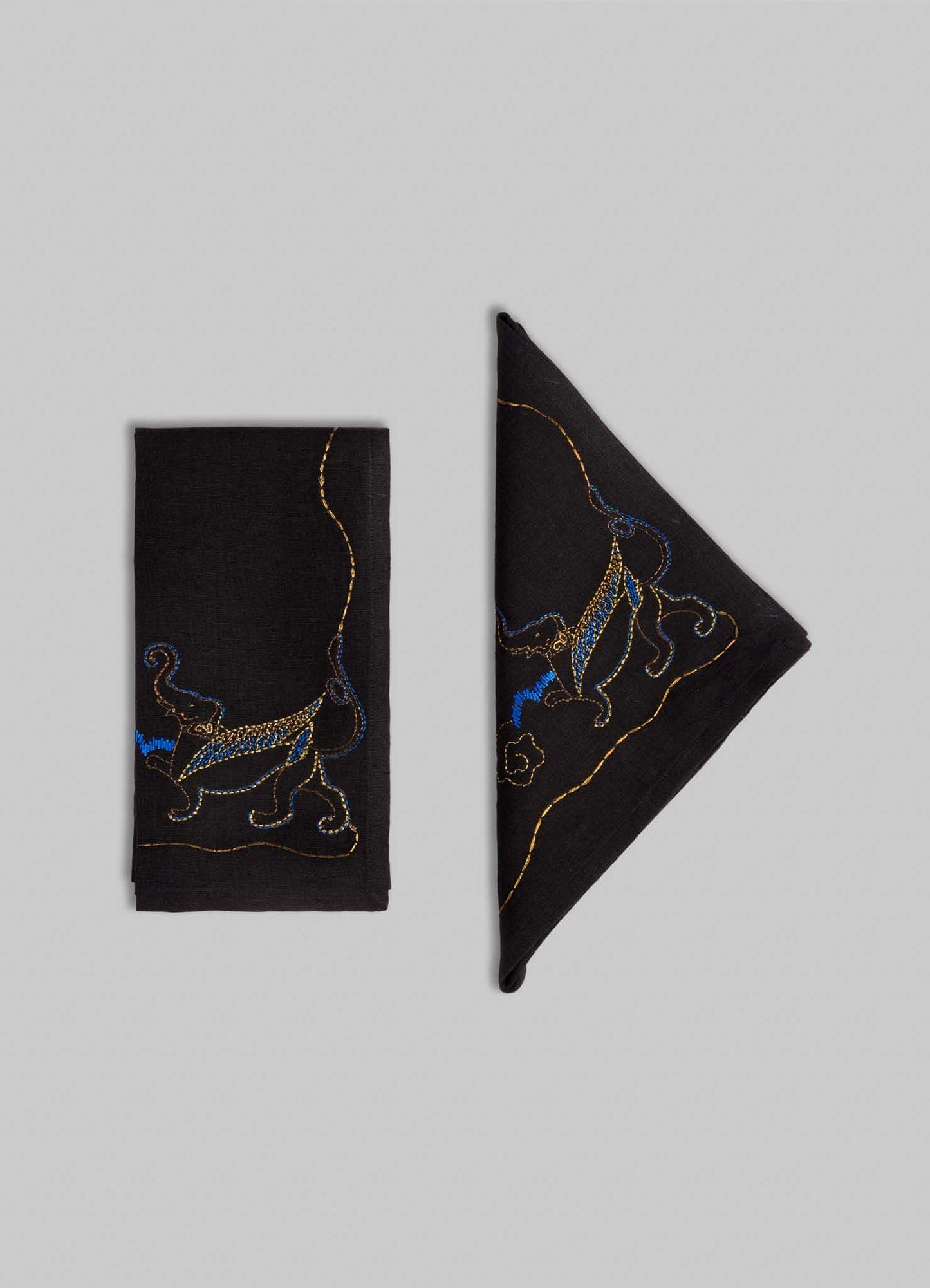 Two Viya Gajasimha Hand Embroidered Black & Gold Napkins, inspired by the ancient Indian mythical guardian, are shown—one folded rectangularly, the other triangularly—on a light gray background.