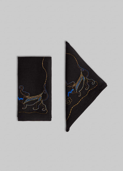 Two Viya Gajasimha Hand Embroidered Black & Gold Napkins, inspired by the ancient Indian mythical guardian, are shown—one folded rectangularly, the other triangularly—on a light gray background.