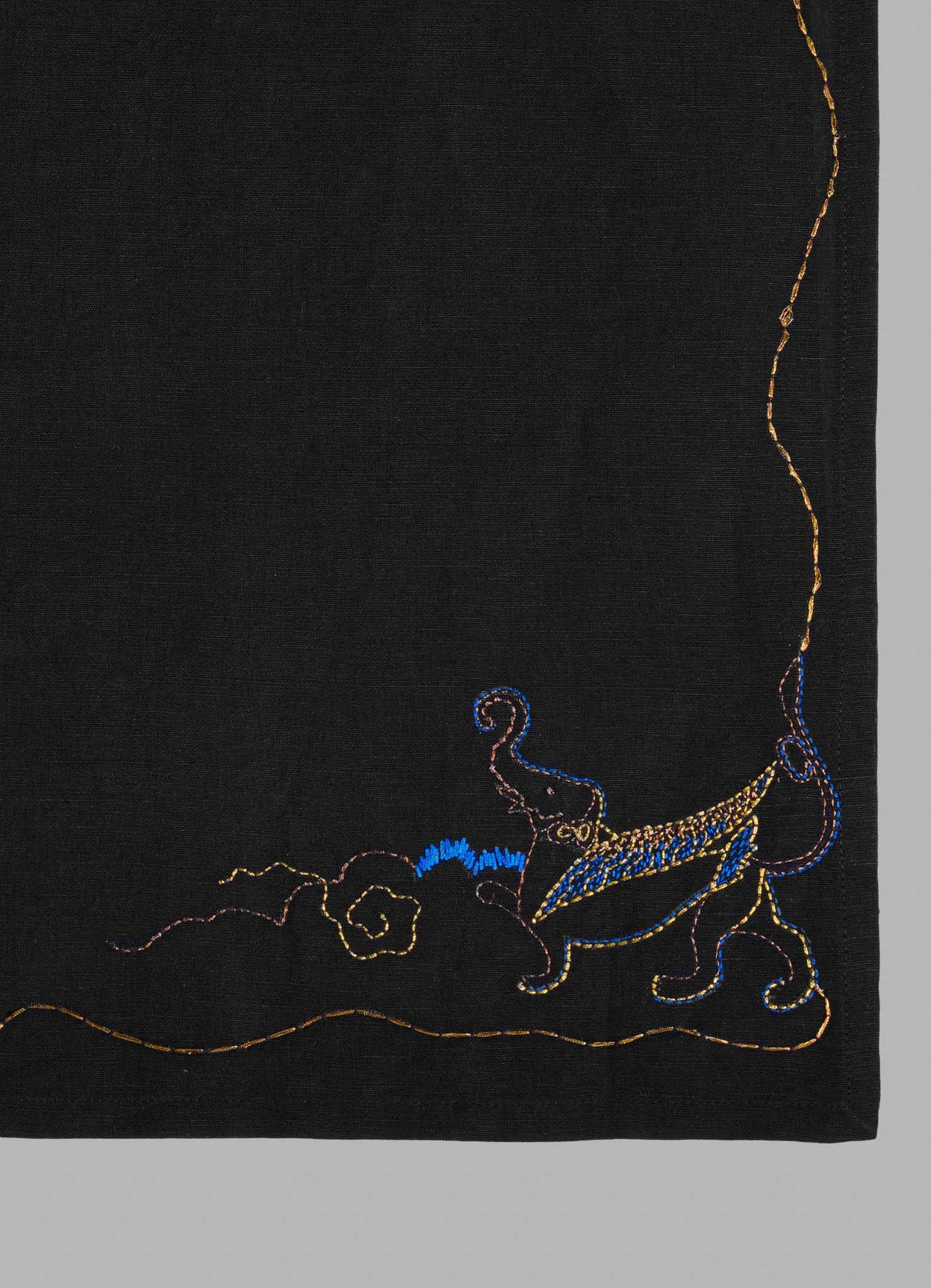 The Viya Gajasimha Hand Embroidered Black & Gold Napkin features a minimalist, colorful Gajasimha motif in blue and yellow on black textile, with golden curved lines and a border, near the lower right corner.