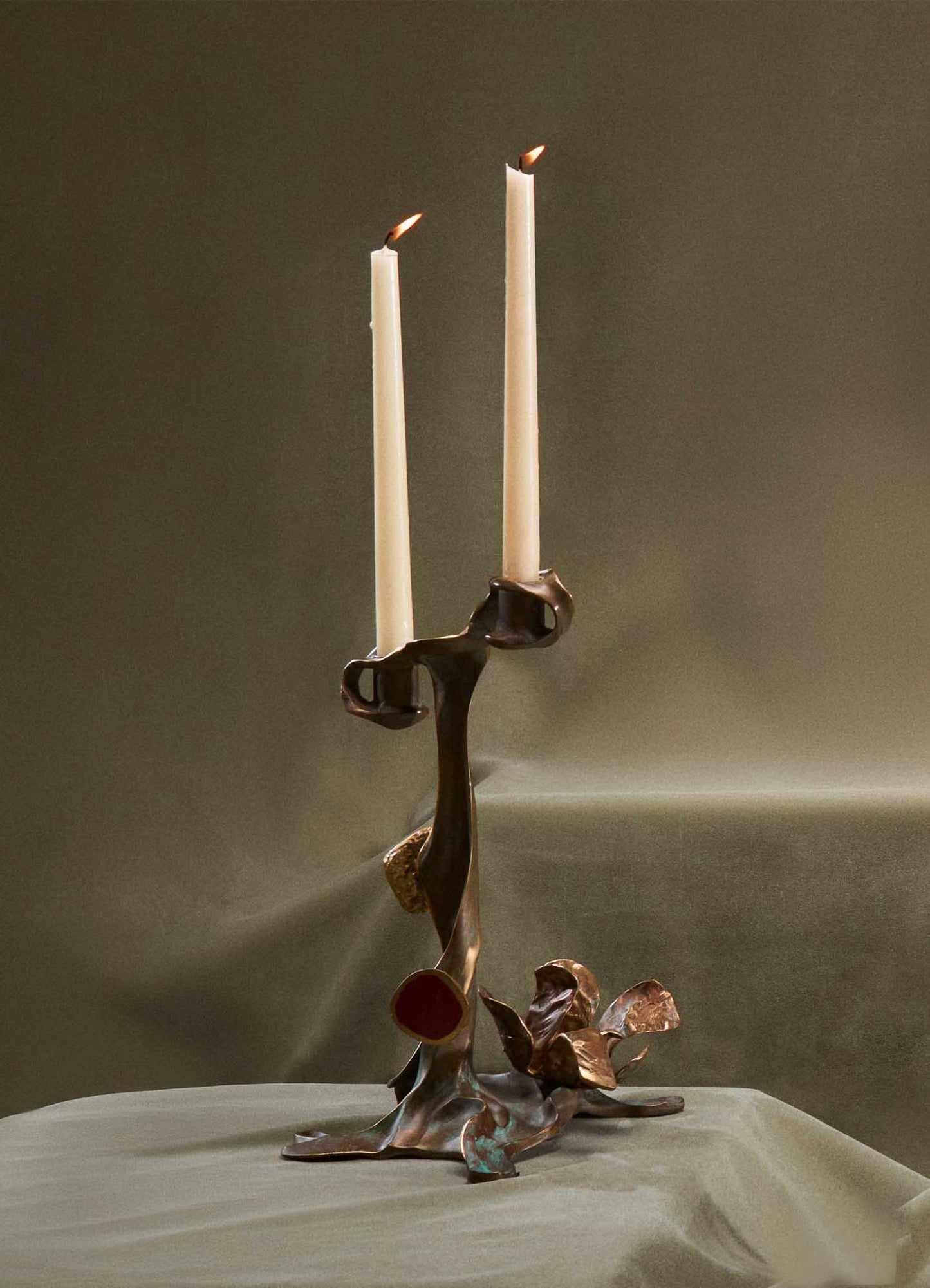 The VIYA Glory Lily Candle Holder Small features sculptural brass with abstract leaves, red and blue accents, and organic forms. It holds two white taper candles, blending tradition with contemporary design against a neutral background.