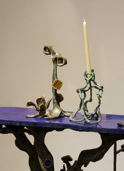 The VIYA Glory Lily Candle Holder Small features an artistic, abstract brass design with sculptural, twisted branch shapes. Displayed on a blue tabletop with a white candle, the contemporary piece stands out against a plain beige background.