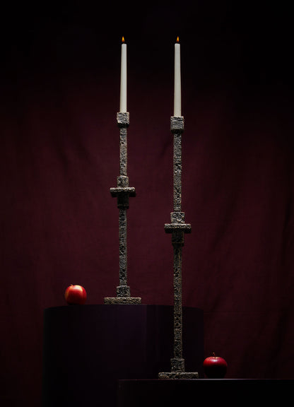 Two tall lit candles in VIYA Tiruppur Totem Candle Holder Large stand on dark pedestals, each with a red apple. They’re set against handwoven fabrics and a deep burgundy background for a dimly lit, dramatic effect.
