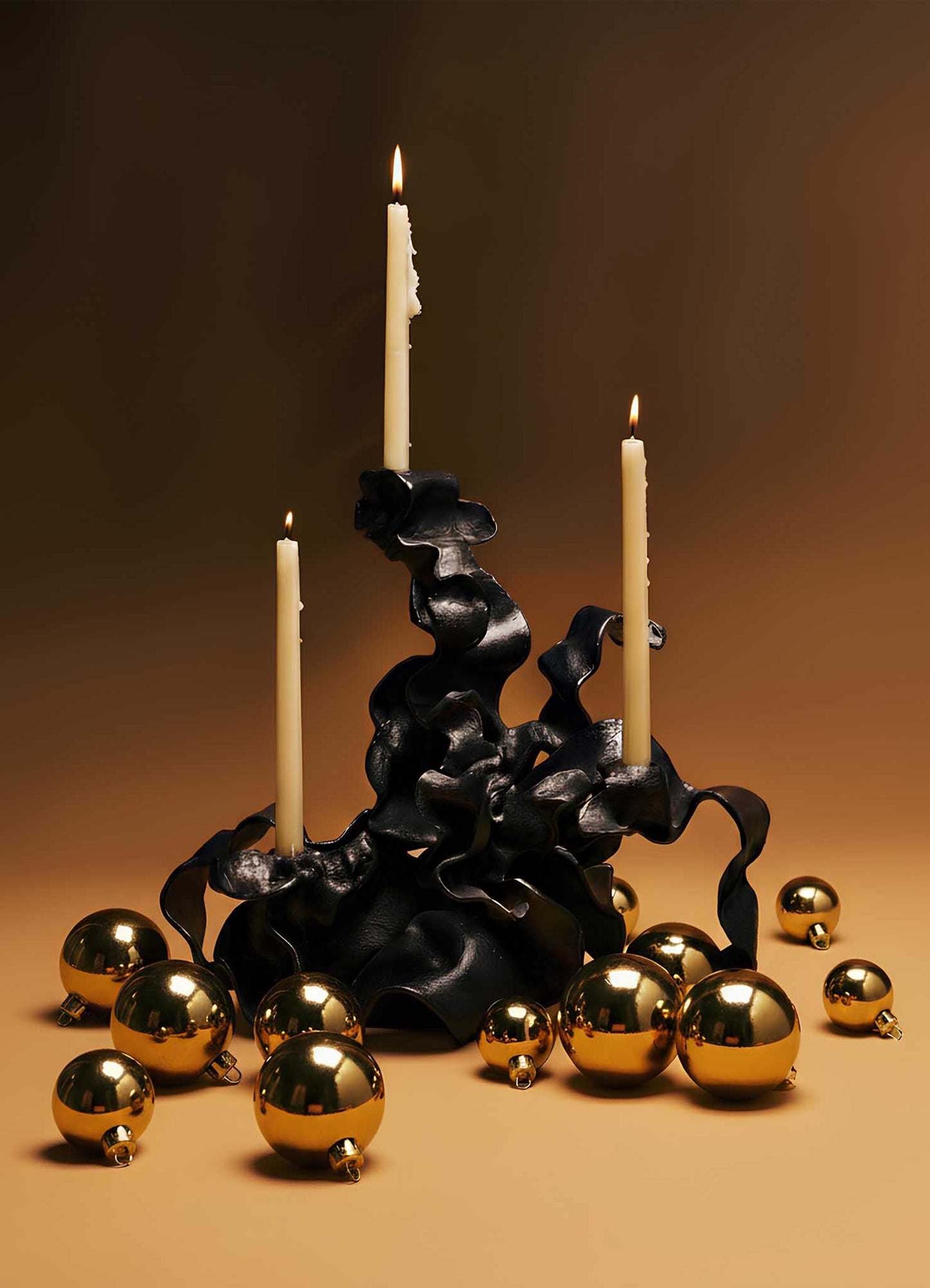 The Hundred Petal Rose Bronze Candelabra by VIYA features three ivory candles in a sculptural black holder, accented with gold ornaments on a warm brown backdrop—two candles lit, one unlit—for elegant home decor.