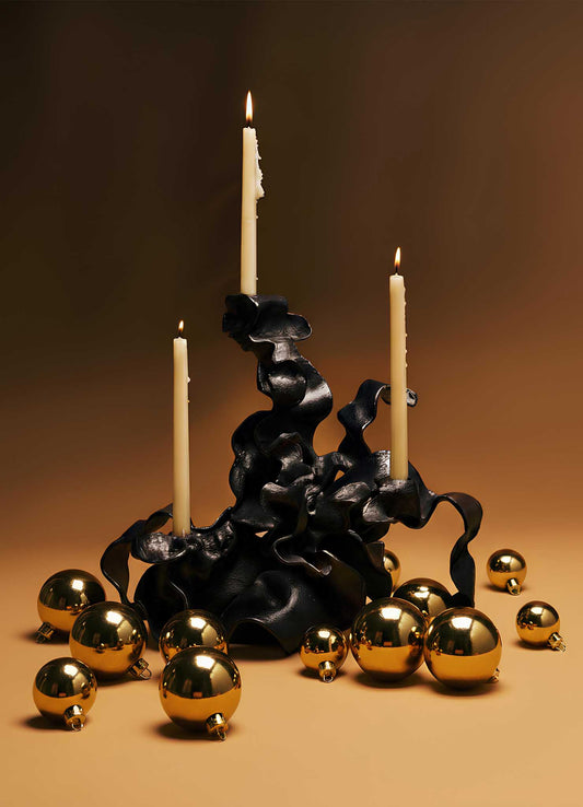 The Hundred Petal Rose Bronze Candelabra by VIYA features three ivory candles in a sculptural black holder, accented with gold ornaments on a warm brown backdrop—two candles lit, one unlit—for elegant home decor.