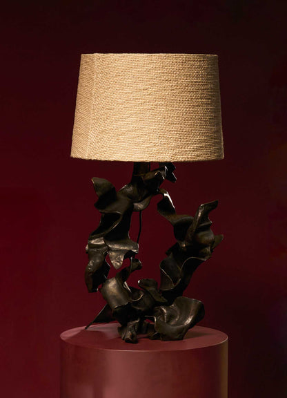 Decorative table lamp with a base featuring abstract, sculptural metal leaves in a dark, brushed finish, topped with a beige fabric lampshade.