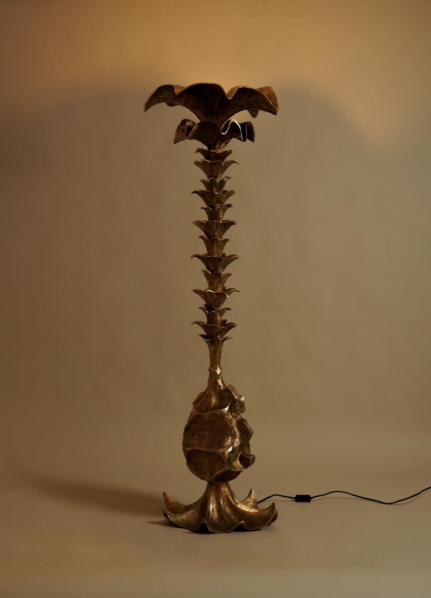 The VIYA Indian Vessel Finial is a tall, sculptural floor lamp with a metallic finish inspired by Indian decor. Featuring a textured trunk and leafy, palm tree-like elements, it has a wide flower-shaped top and a black power cord at its base.