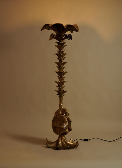 The VIYA Indian Vessel Finial is a tall, sculptural floor lamp with a metallic finish inspired by Indian decor. Featuring a textured trunk and leafy, palm tree-like elements, it has a wide flower-shaped top and a black power cord at its base.