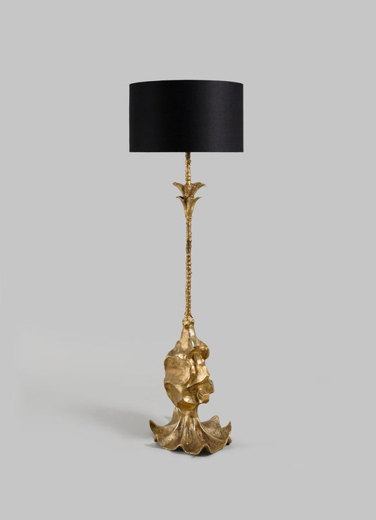 The VIYA Indian Vessel Floor Lamp features a tall, sculpted gold metallic base inspired by brass vessels and abstract forms, with a slender stem and large cylindrical black lampshade, displayed against a plain gray background.