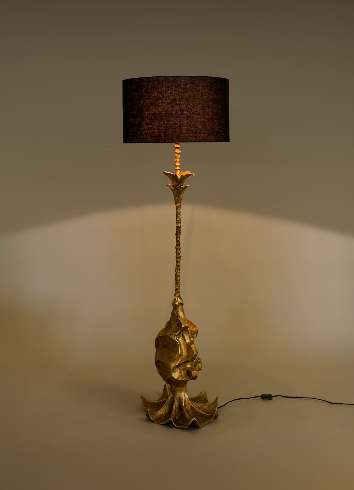 The VIYA Indian Vessel Floor Lamp features a sculpted gold base and stem inspired by brass vessels, topped with a black cylindrical shade and standing on a plain surface with a visible power cord.