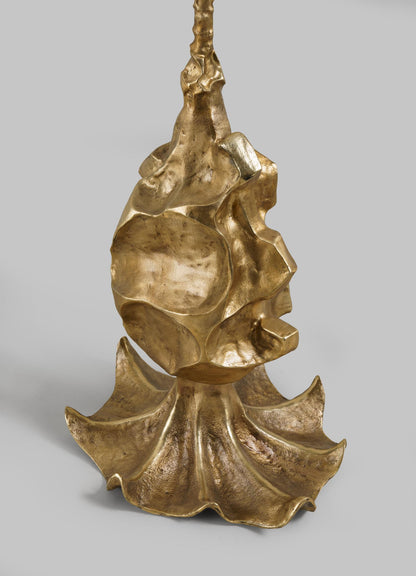 The VIYA Indian Vessel Floor Lamp features a sculpted metallic design with a shiny gold finish, abstract curves inspired by lota or surahi brass vessels, and stands upright on a wavy, textured base reminiscent of a stylized leaf or flower.