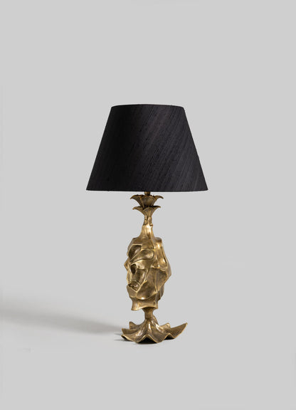 The VIYA Indian Vessel Table Lamp features a sculpted gold base inspired by traditional brass vessels and abstract forms, paired with a black conical shade and displayed against a plain light gray background.
