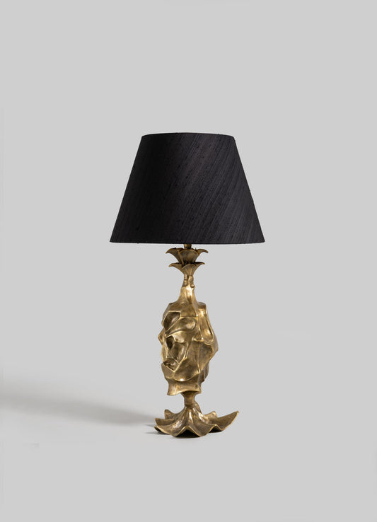 The VIYA Indian Vessel Table Lamp features a sculpted gold base inspired by traditional brass vessels and abstract forms, paired with a black conical shade and displayed against a plain light gray background.