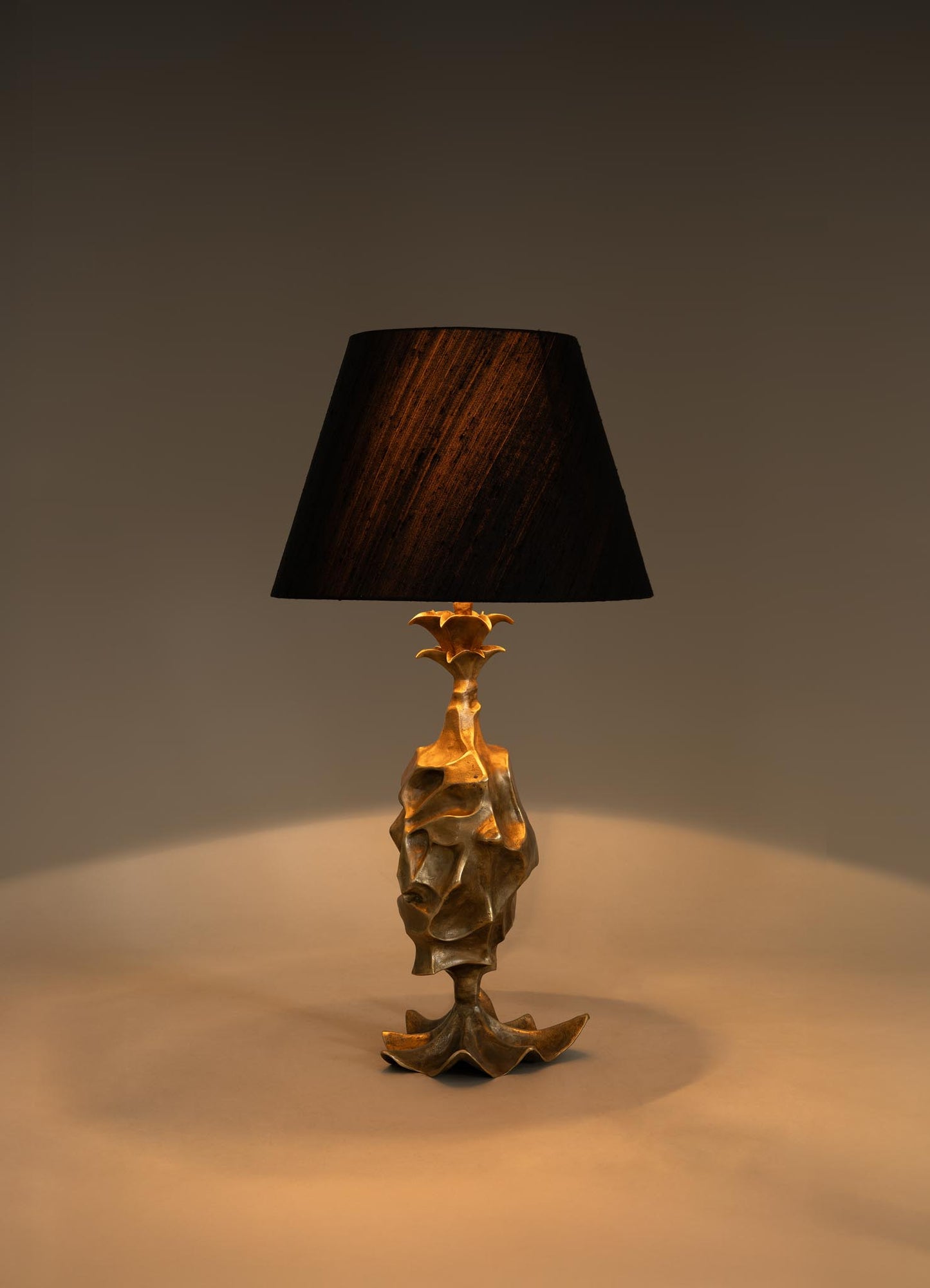 The VIYA Indian Vessel Table Lamp features a sculptural gold base inspired by traditional brass vessels and a dark, subtly reflective lampshade, elegantly displayed on a neutral surface against a plain background.