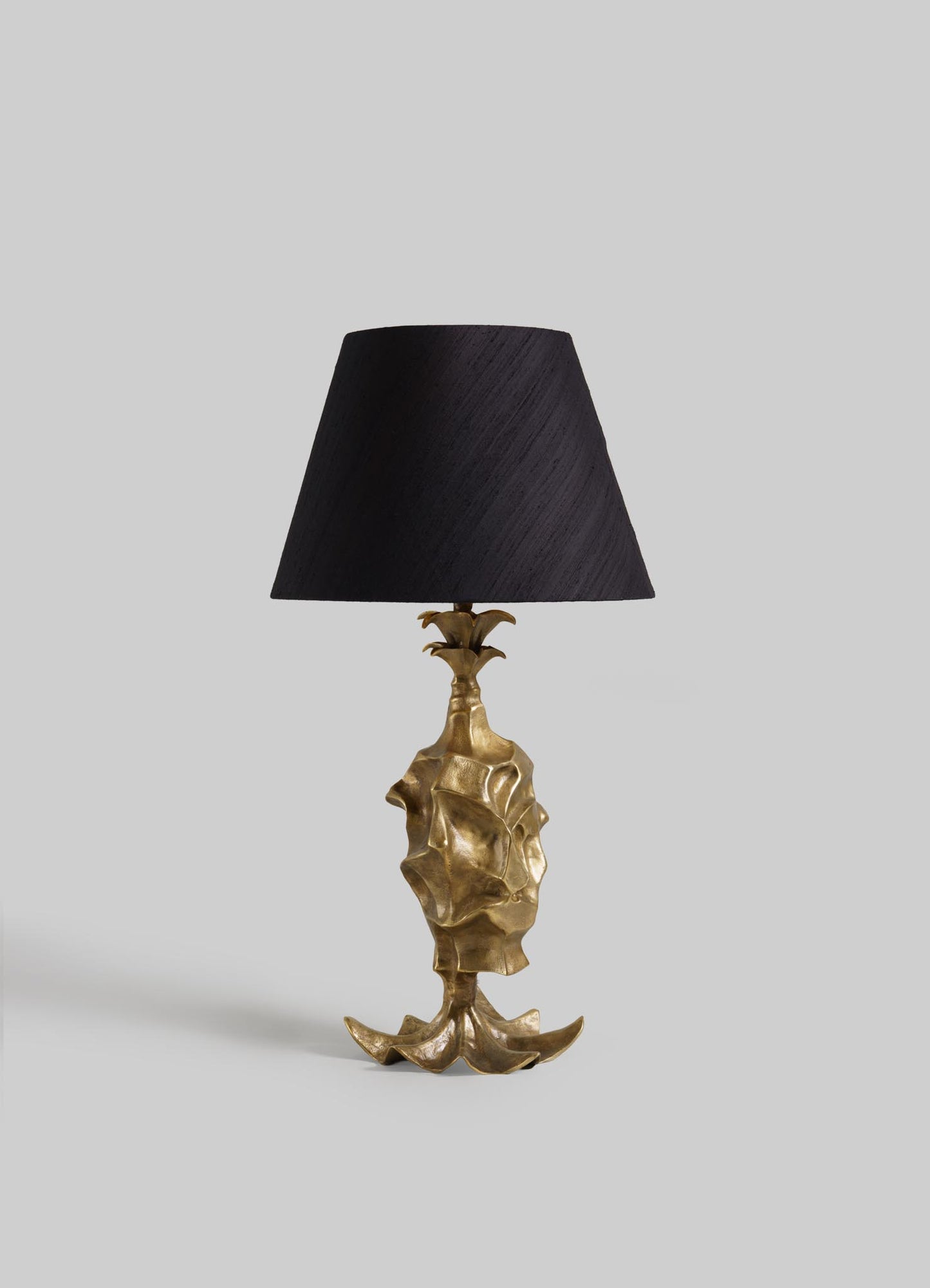 The VIYA Indian Vessel Table Lamp features a sculpted base inspired by traditional brass surahi vessels and a wide black lampshade, shown against a plain light gray background.