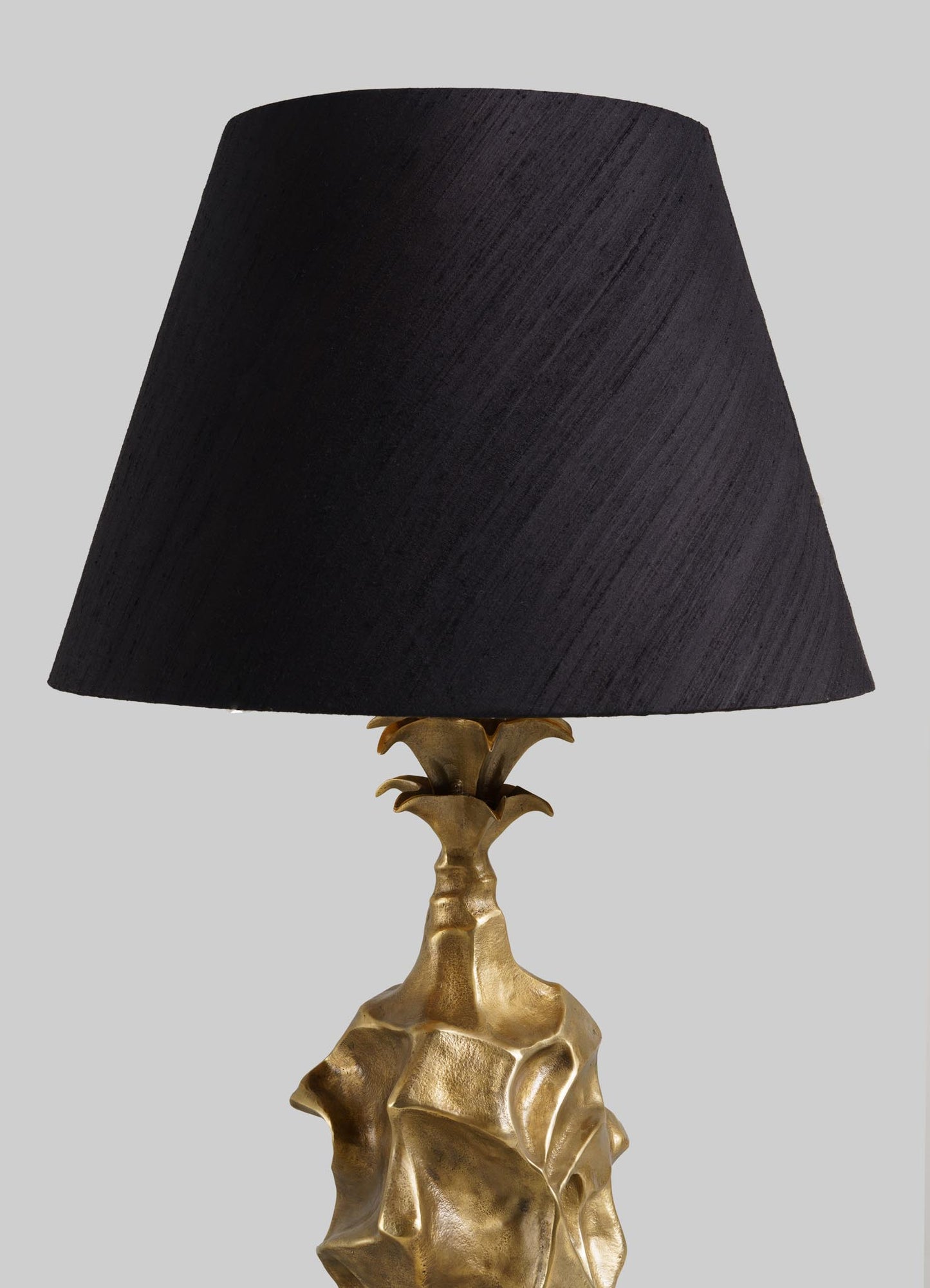 The VIYA Indian Vessel Table Lamp features a sculpted gold base inspired by abstract brass vessels and a large black lampshade, displayed against a plain light gray background.