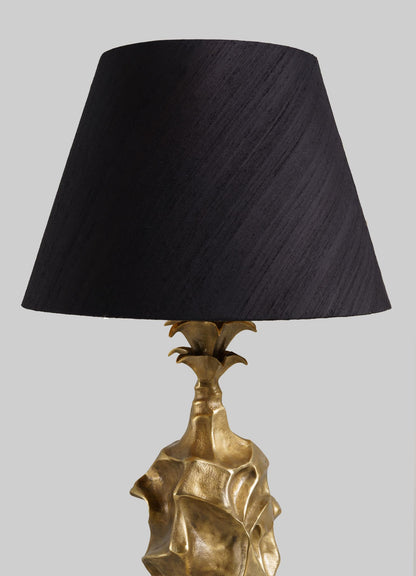 The VIYA Indian Vessel Table Lamp features a sculpted gold base inspired by abstract brass vessels and a large black lampshade, displayed against a plain light gray background.